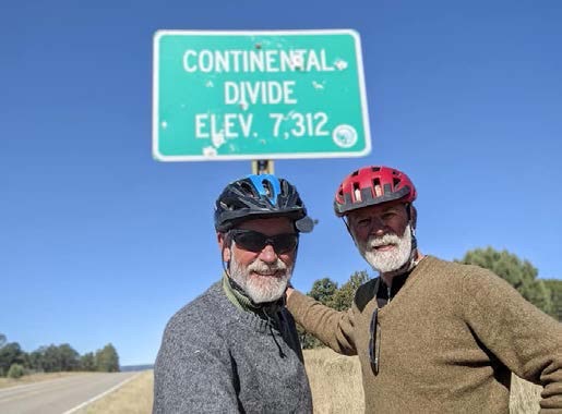 ALT TEXT PHOTO: Ferdinand Lauffer standing side-by-side and his arm around his buddy both wearing bike helmets, sweaters and sporting long wooly white beards. Behind them, is blue skies and a sign clearly stating Continental Divide - Elevation 7,321 feet.
