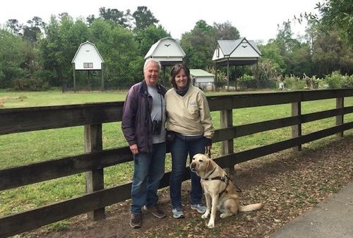 ALT TEXT PHOTO: Martha Steele with her husband, Bob Stymeist, and her guide dog, Alvin, standing outside with a green field and special structures for birding behind them.