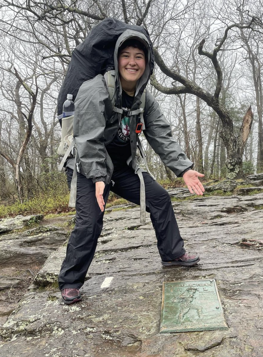 ALT TEXT PHOTO: Lo Makechnie's first day on the Appalachian Trail in Georgia with bare trees behind them, smiling at the camera wearing their rain jacket with a large-trail backpack and gesturing to a placard that says, Appalachian Trail, bolted into the large flat-faced rock that Lo is standing on.