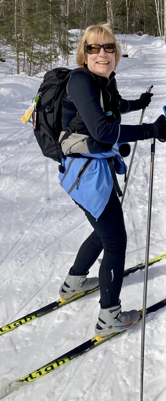 ALT TEXT PHOTO: Diane Manganaro, adorned in winter gear, a backpack, and skis radiates a warm smile while gracefully striking a pose with her ski poles in hand. Behind her, a picturesque winter wonderland unfolds, with snow-draped ground and a forest of trees setting a wintry backdrop.