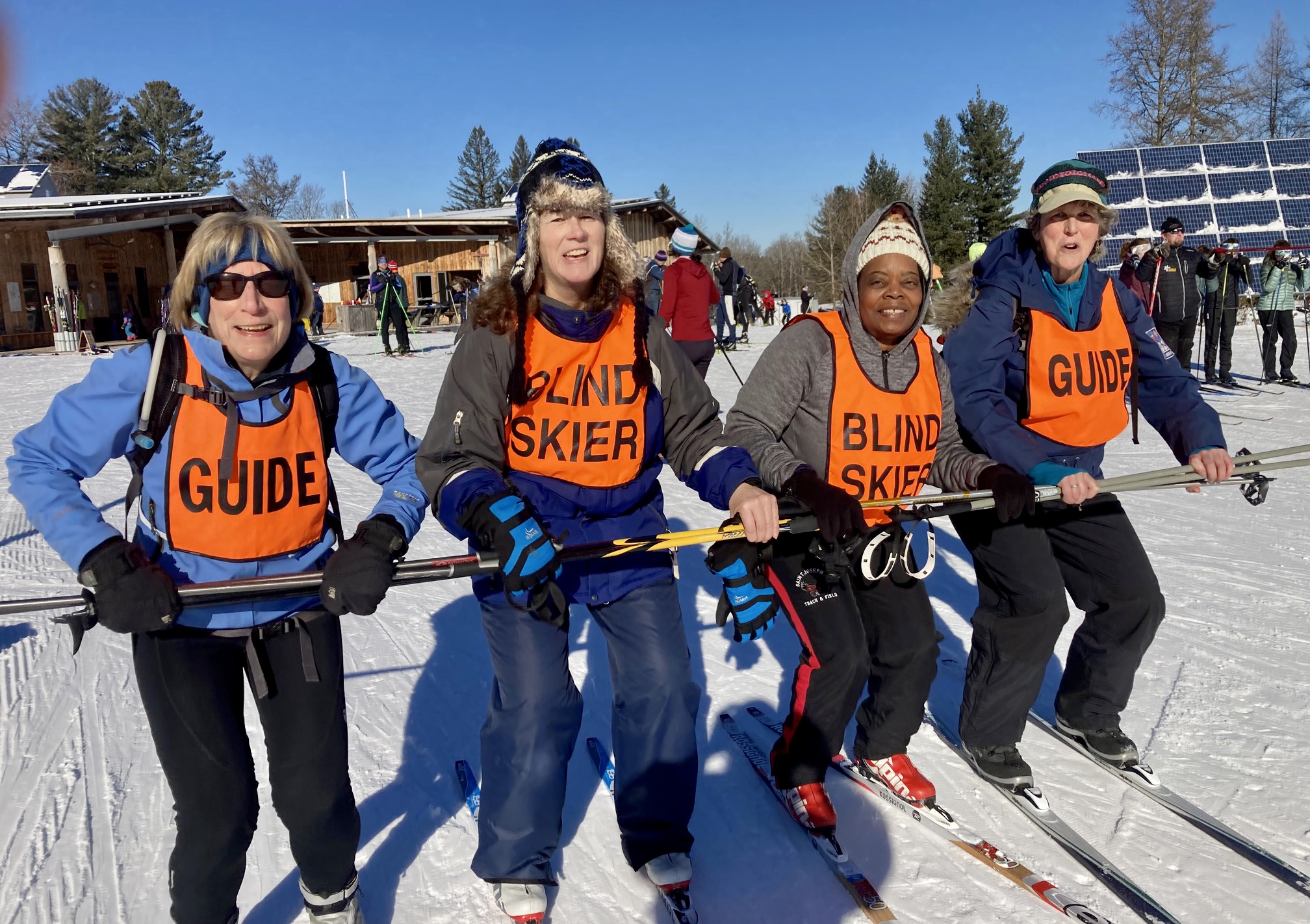 ALT TEXT PHOTO: A quartet of smiling skiers, wearing winter attire and skis, clutch interlinking ski poles held out in front of them. To the left stands Diane Manganaro, wearing a vibrant orange ski bib bearing the word 